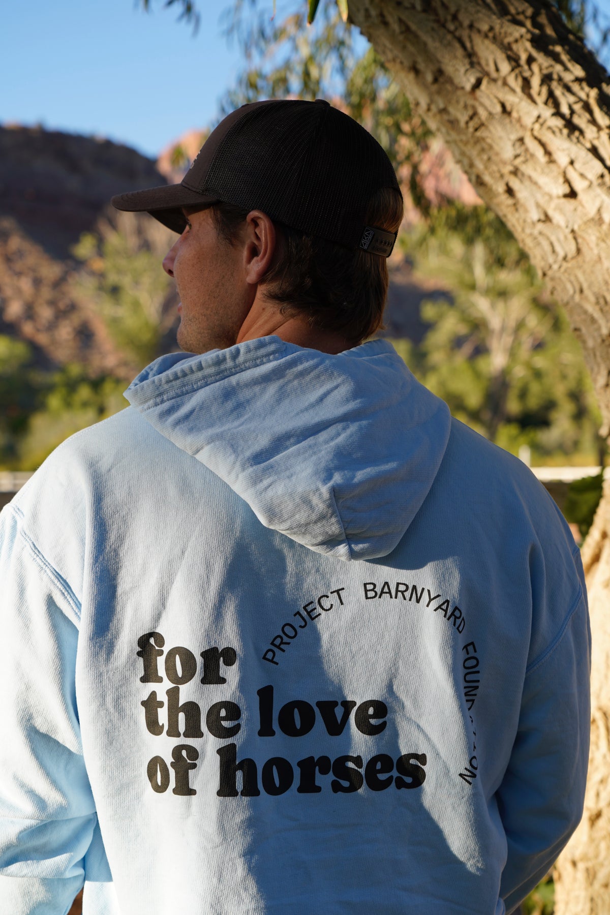A man wearing a light blue hoodie with black text on the back reading 'for the love of horses' and 'PROJECT BARNYARD CLOTHING', photographed outdoors near a tree with hills in the background.