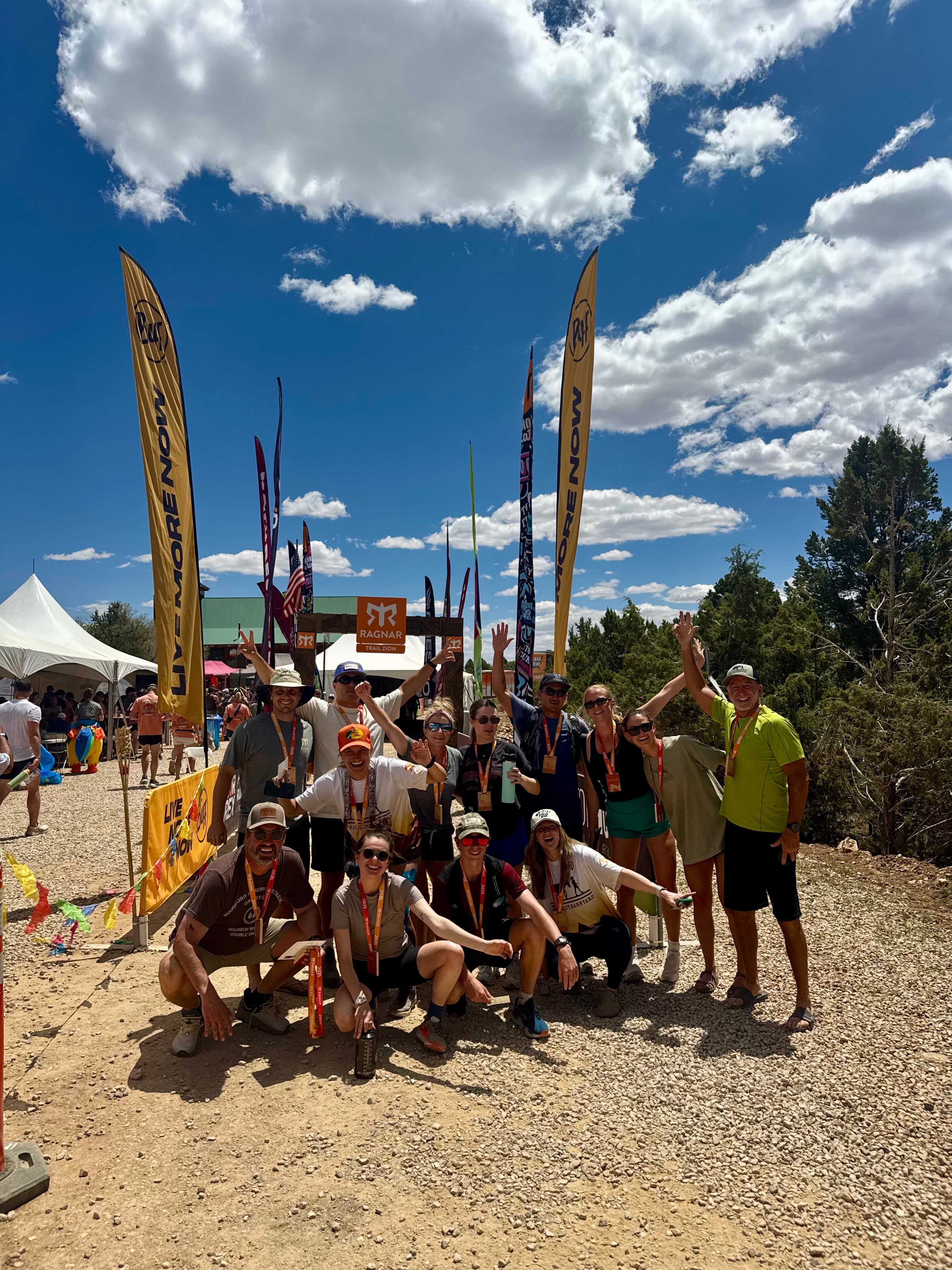 A group of trail runners posing together outdoors at a race or event, with tall branded feather flags, tents, trees, and a blue sky with clouds in the background.