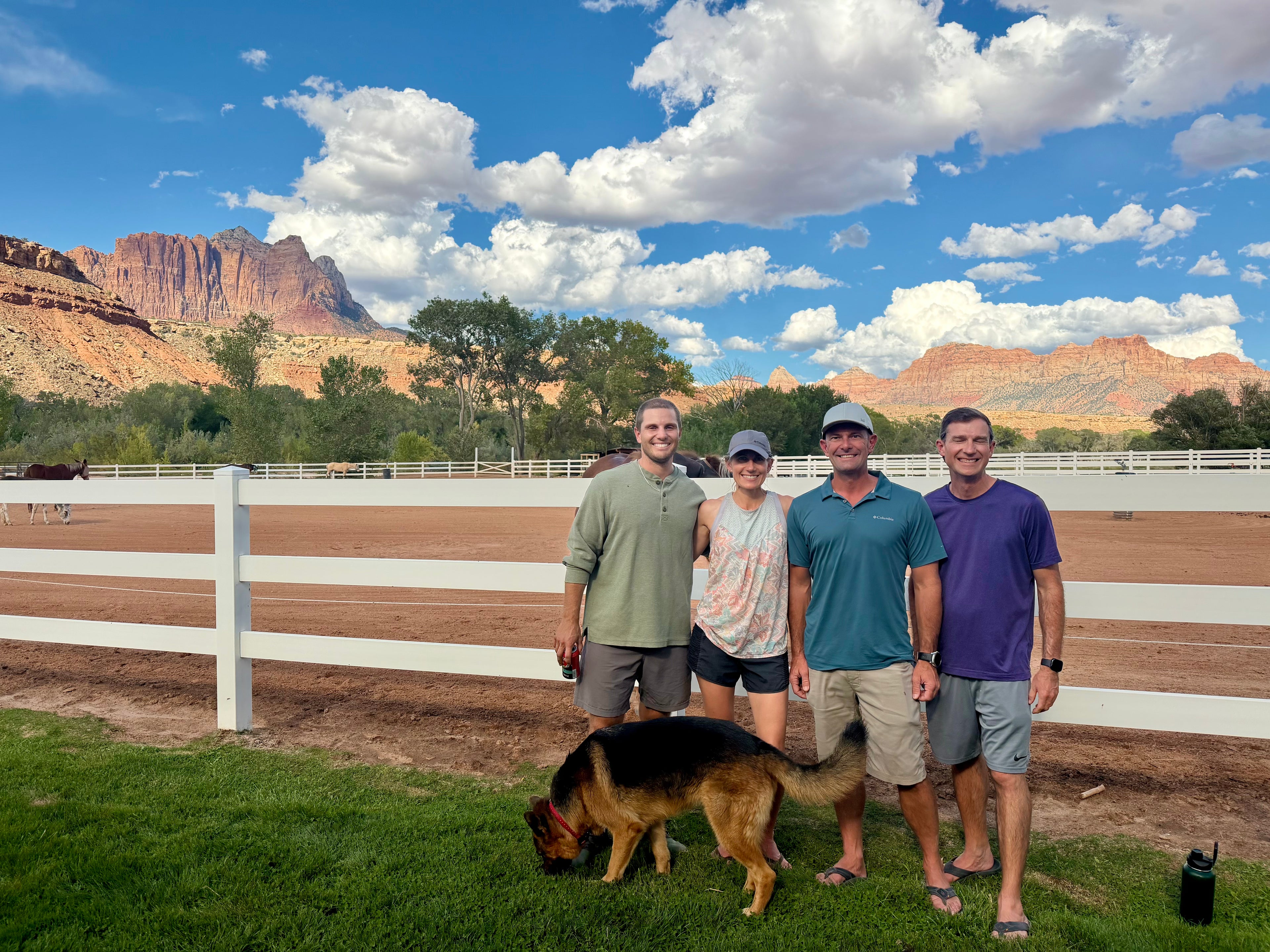 Four men standing outdoors on a grassy field in front of a white fence and dirt riding arena, with red rock mountains and a blue sky with clouds behind them. A German shepherd is in the foreground near their feet.