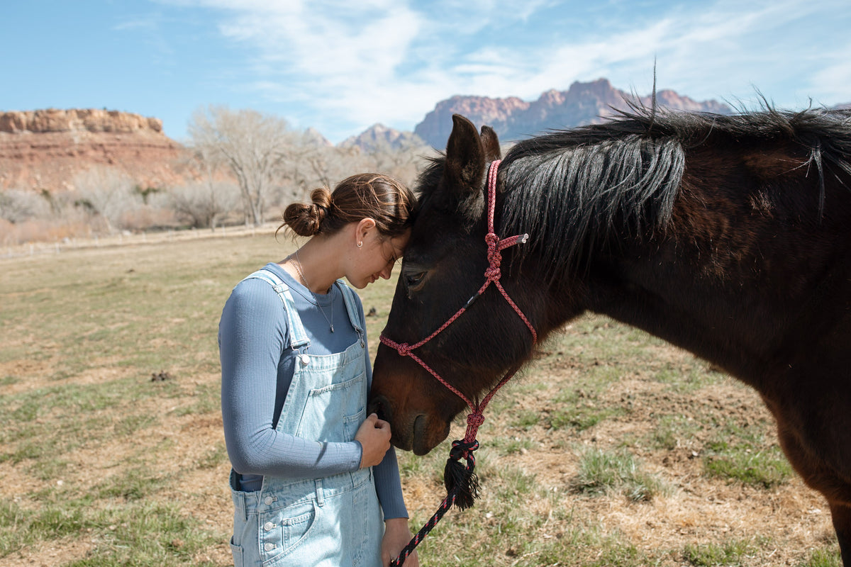 A woman in light blue denim overalls and a gray long-sleeve shirt stands in a grassy field next to a dark brown horse wearing a red rope halter, with red rock mountains and a blue sky in the background.