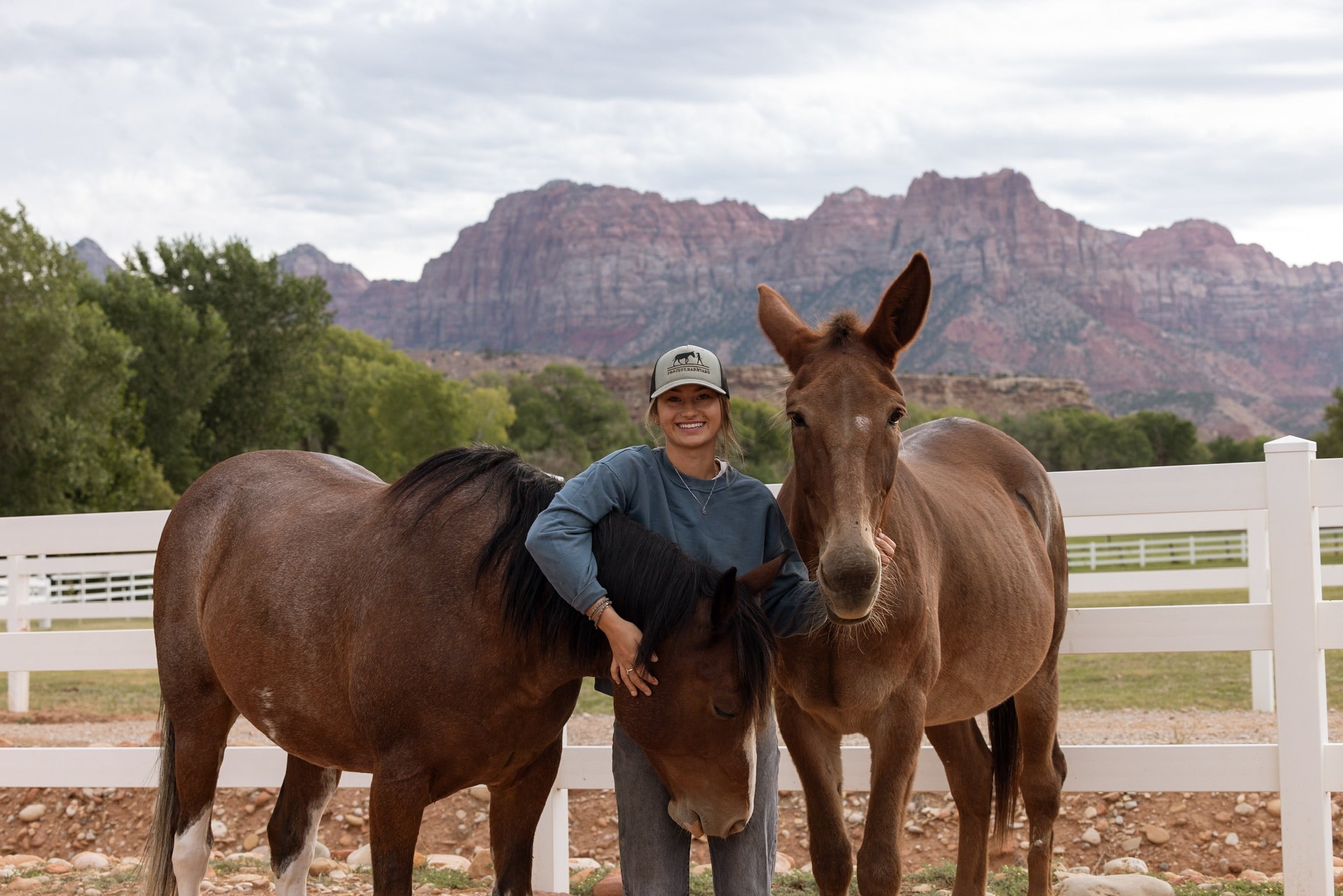 A smiling woman stands outdoors between two brown horses in a fenced pasture, with white fencing and large red rock mountains in the background under a cloudy sky.